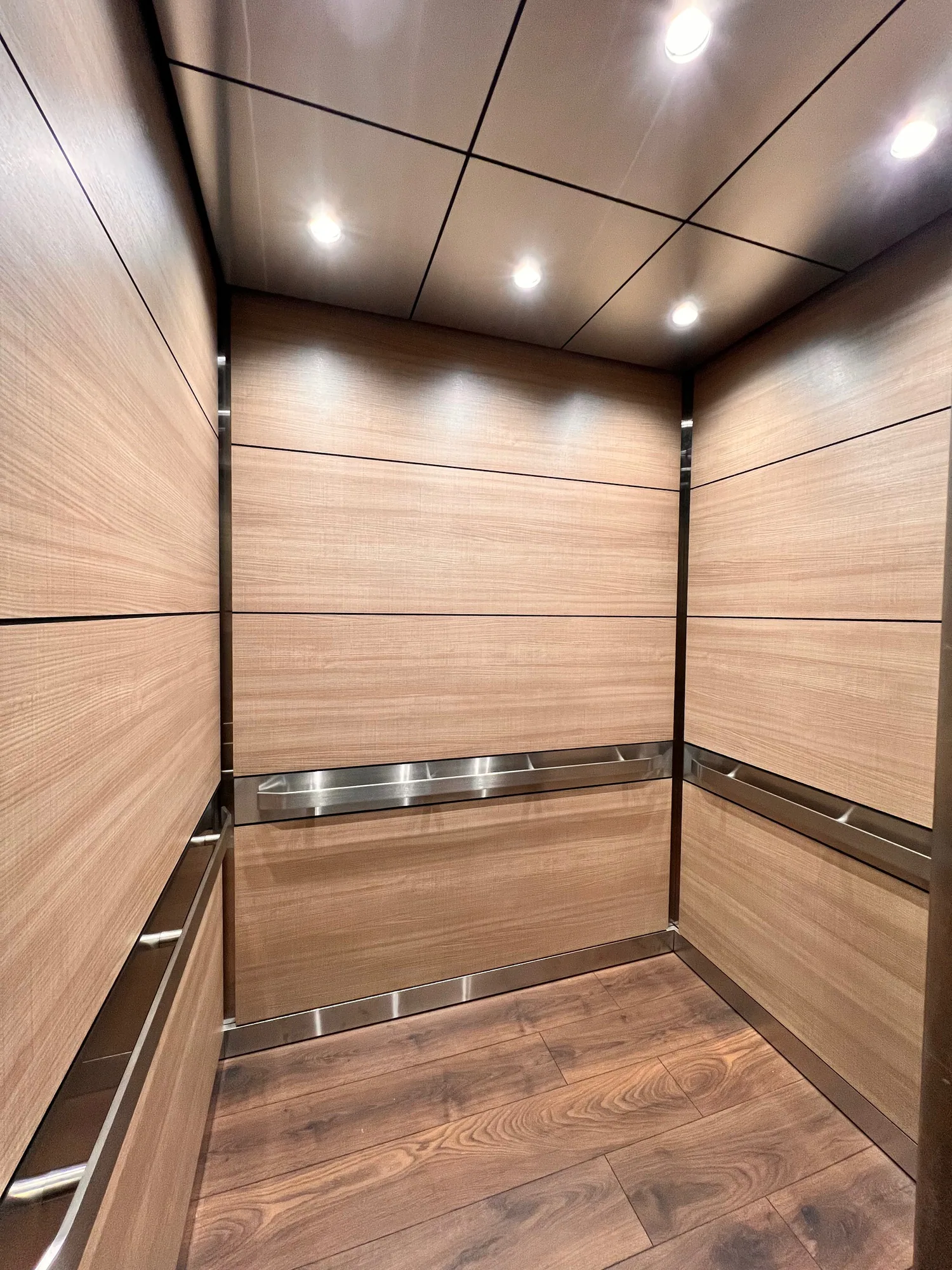 Warm woodgrain elevator interior with stainless handrails and illuminated ceiling panels